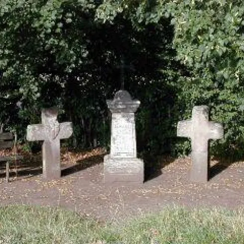 Ein kleiner Friedhof mit drei Grabkreuzen, umgeben von Bäumen. Ein Holzbank ist ebenfalls sichtbar.
