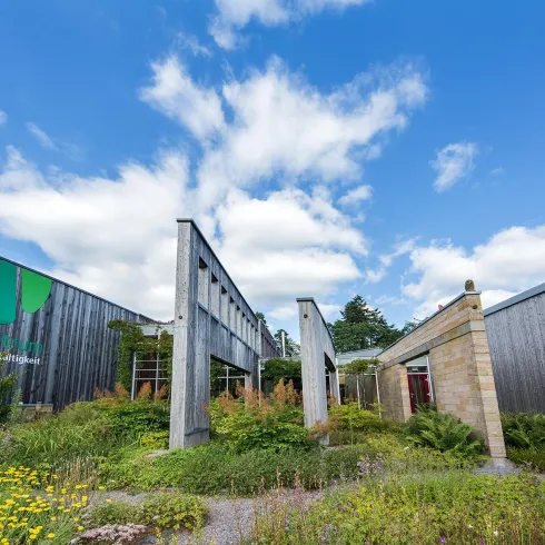 Ein modernes, architektonisches Gebäude mit einer Mischung aus Holz und Stein. Es ist von grüner Vegetation umgeben und der Himmel ist klar und blau.