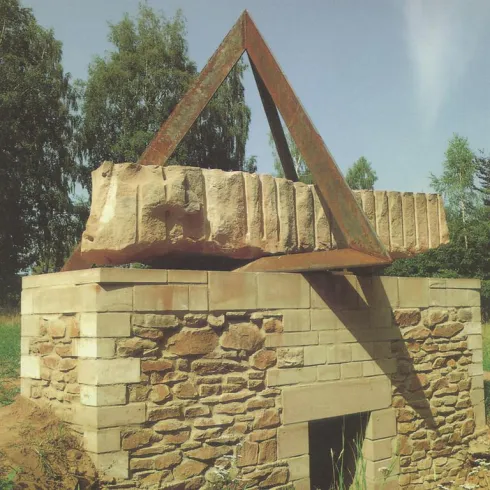 Ein moderner Steinskulptur besteht aus einem quadratischen Sockel aus Naturstein und einem dreieckigen Metallrahmen, der einen großen Stein blockiert. Die Umgebung ist grün mit Bäumen im Hintergrund.