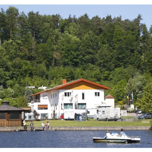 Ein ruhiger See mit einem Boot und einer weißen Unterkunft am Ufer. Dahinter sind Bäume und eine grüne Landschaft zu sehen.