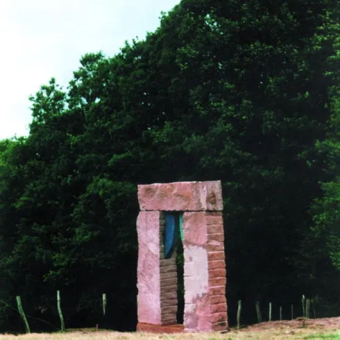 Ein Steindenkmal steht auf einer Wiese, umgeben von Bäumen. Das Monument besteht aus großen Steinblöcken mit einer Öffnung in der Mitte.
