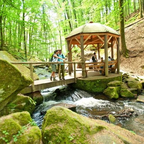 Eine idyllische Waldlandschaft mit einem kleinen Pavillon über einem klaren Bach. Menschen genießen die Natur und die Umgebung ist von grünen Bäumen und Felsen umgeben.