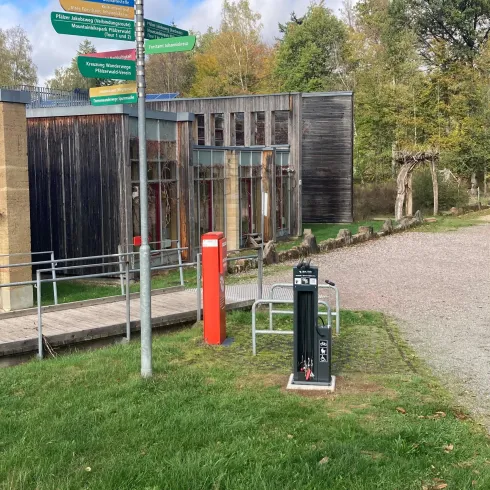 Eine moderne Gebäudeansicht mit einem Wegweiser und einer roten Telefonzelle im Vordergrund. Im Hintergrund sind Bäume und ein blauer Himmel zu sehen.