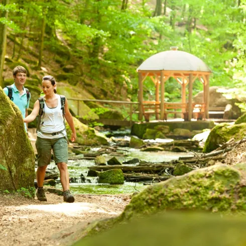 Zwei Wanderer spazieren auf einem Weg durch einen grünen Wald. Im Hintergrund ist ein Pavillon und ein kleiner Bach sichtbar.