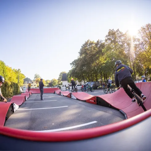 Eine lebhafte Actionszene mit Radfahrern in einer Pumptrack-Anlage. Die Umgebung ist grün mit Bäumen und es scheint sonnig zu sein.