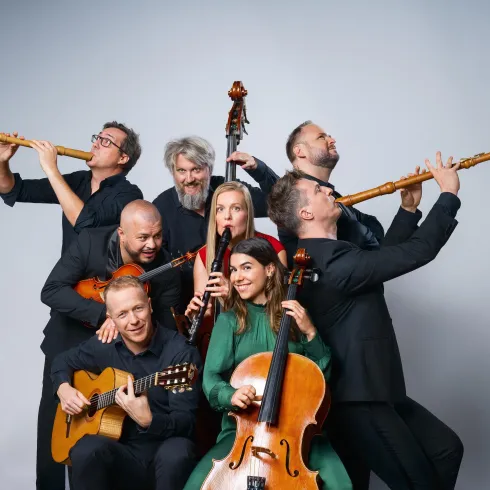 Eine Gruppe von Musikern mit verschiedenen Instrumenten steht zusammen. Sie lächeln und posieren fröhlich in einem neutralen Hintergrund.