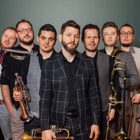 Eine Gruppe von acht Musikern steht zusammen und hält verschiedene Blechblasinstrumente. Sie tragen elegante Kleidung und lächeln in die Kamera.