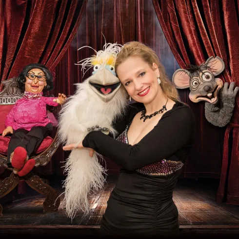 Eine Frau steht auf der Bühne und hält eine Puppenfigur. Im Hintergrund sind zwei weitere Puppen in einem Bühnenarrangement zu sehen.