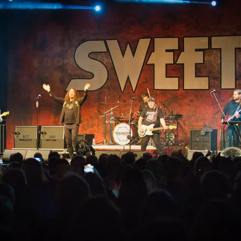 Eine Rockband tritt auf einer Bühne vor einem begeisterten Publikum auf. Im Hintergrund ist das große Logo "SWEET" zu sehen.