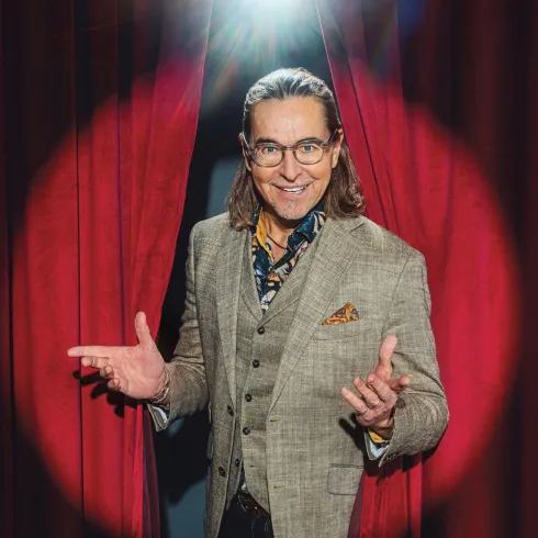 Ein Mann mit langen Haaren steht im Spotlight und lächelt. Er trägt einen eleganten Anzug und steht vor einem roten Vorhang.