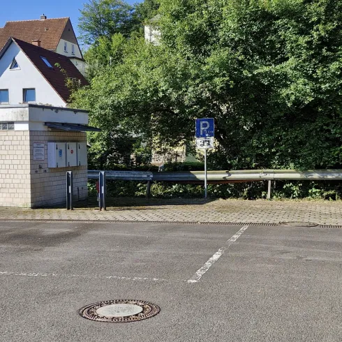Eine ruhige Straße mit einem Parkplatzschild und angrenzendem Grünbereich. Im Hintergrund sind Häuser und Bäume sichtbar.