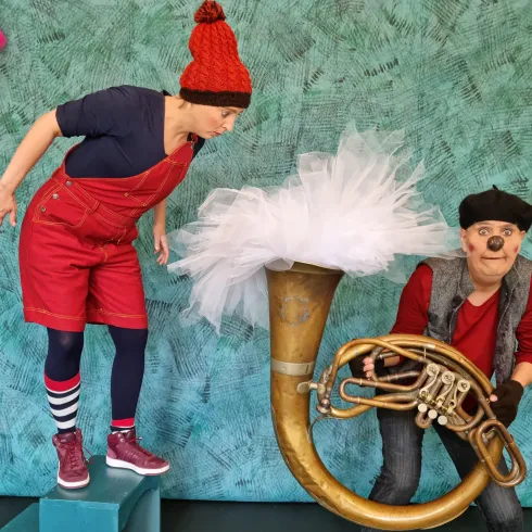 Zwei Darsteller in bunten Kostümen stehen auf einer Bühne. Einer schaut neugierig auf ein großes Holzblasinstrument, während der andere es hält.