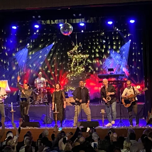 Eine lebhafte Band spielt auf einer Bühne vor einem begeisterten Publikum. Farbiges Licht und Dekorationen sorgen für eine festliche Atmosphäre.