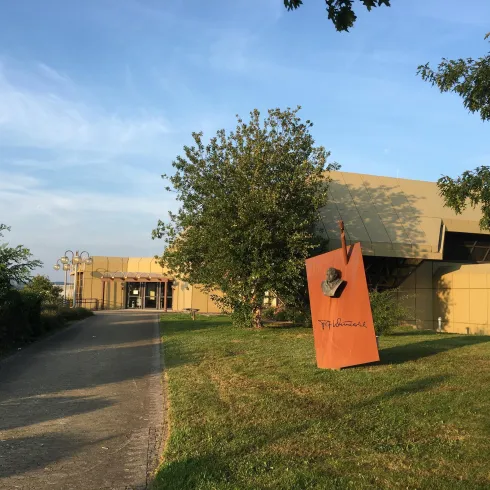 Ein modernes Gebäude umgeben von Grünflächen und Bäumen. Der Weg führt zur Eingangstür des Gebäudes.