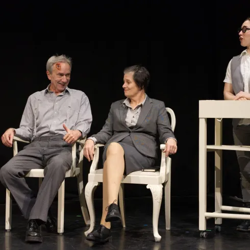 Drei Personen auf einer Bühne in einem Theater. Zwei Personen sitzen auf Stühlen, während eine dritte Person an einem Tisch steht.