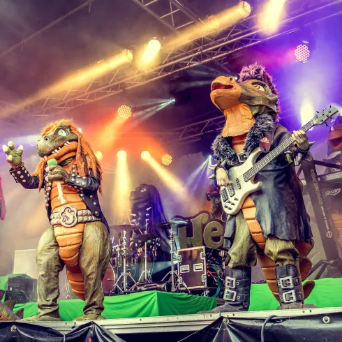 Eine Band in Dinosaurier-Kostümen spielt auf einer Bühne. Bunte Lichter und Nebel schaffen eine festliche Atmosphäre.