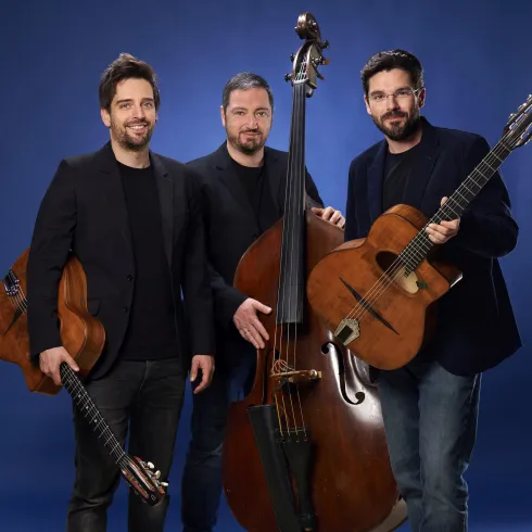 Drei Musiker stehen in eleganter Kleidung vor einem blauen Hintergrund. Einer hält eine Gitarre, ein anderer einen Kontrabass und der dritte eine Gitarre in der Hand.