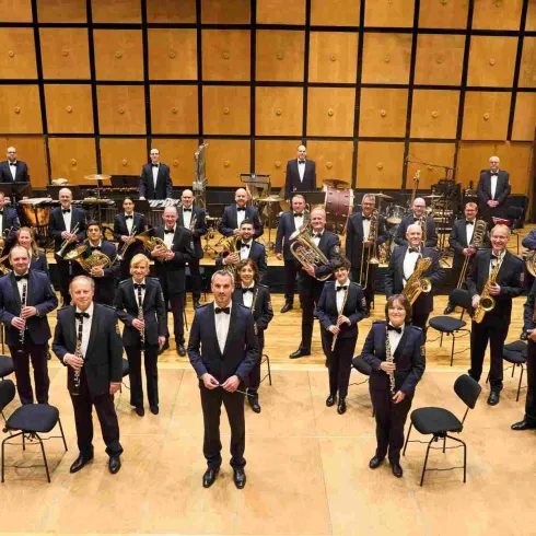 Ein Orchester steht auf der Bühne in eleganter Kleidung. Es besteht aus Musikern, die verschiedene Instrumente spielen und lächeln.