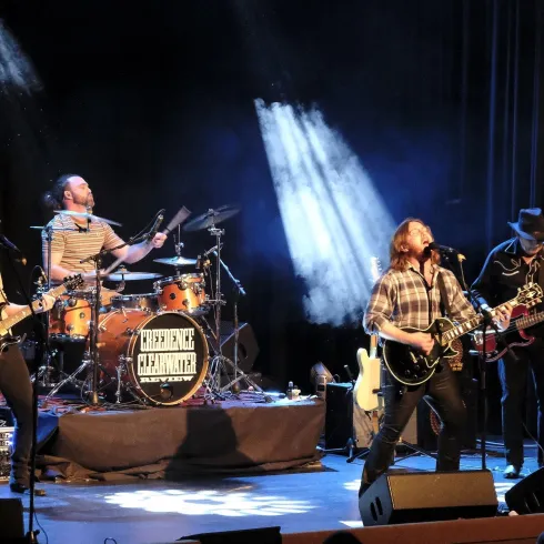 Eine Band spielt auf der Bühne vor einem dunklen Hintergrund. Die Musiker sind leidenschaftlich bei ihrem Auftritt und nutzen verschiedene Instrumente.
