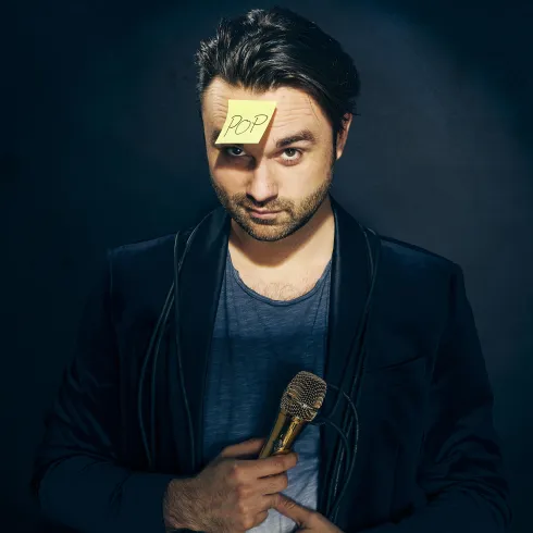 Ein Mann mit einem Mikrofon in der Hand und einem Klebezettel mit der Aufschrift "POP" auf der Stirn. Er trägt ein lässiges Outfit und schaut direkt in die Kamera.