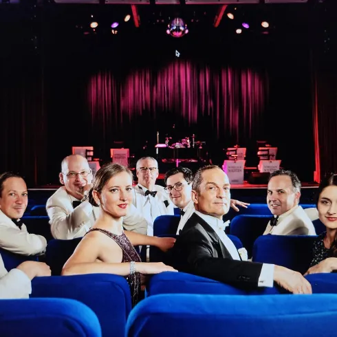 Eine Gruppe von Menschen in eleganter Kleidung sitzt in blauen Stühlen. Im Hintergrund ist eine Bühne mit Lichtern und Musikinstrumenten zu sehen.