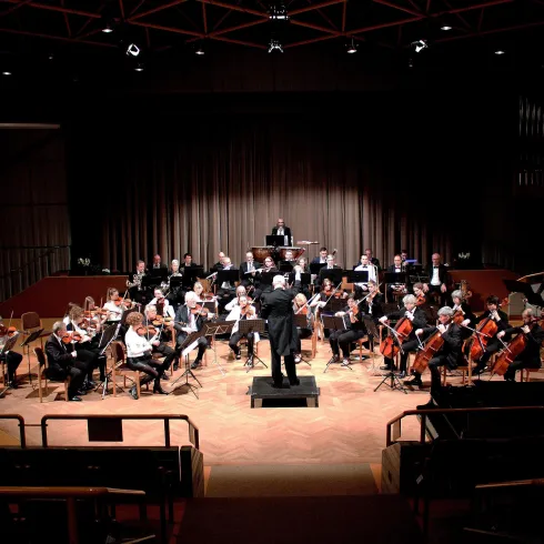 Ein Orchester spielt auf einer Bühne mit einem Dirigenten. Die Musiker sitzen in typischer Anordnung mit verschiedenen Instrumenten.