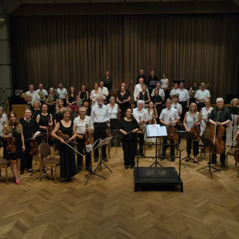 Eine große Gruppe von Menschen steht lächelnd auf einer Bühne. Sie sind Musiker in Konzertkleidung und halten verschiedene Musikinstrumente.