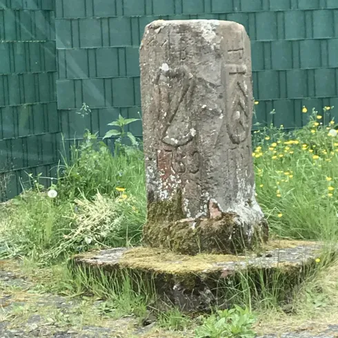 Ein steinerner Obelisk mit Schnitzereien steht im Grünen. Um ihn herum wächst Gras und es gibt kleine Blumen.