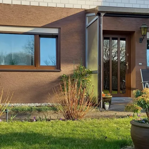 Ein modernes Haus mit brauner Fassade und großen Fenstern. Der Garten ist gepflegt und enthält einige Pflanzen und Sitzgelegenheiten.