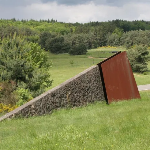 Eine schiefe Skulptur aus Metall und Stein steht in einer grünen Landschaft. Im Hintergrund sind Bäume und ein wolkenverhangener Himmel sichtbar.