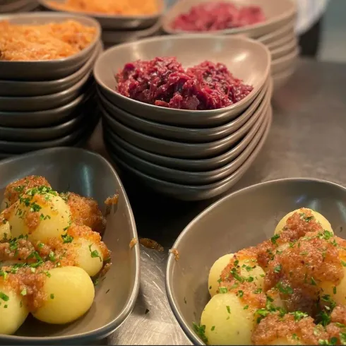 Ein Teller mit kleinen, goldenen Kartoffeln, garniert mit Kräutern. Im Hintergrund stehen mehrere Schalen mit rot und braunem Beilagen.