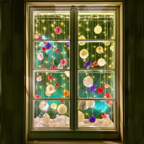 Ein festlich dekoriertes Fenster mit bunten Ornamenten und Lichtern. Im Hintergrund ist ein grüner Weihnachtsbaum zu sehen.