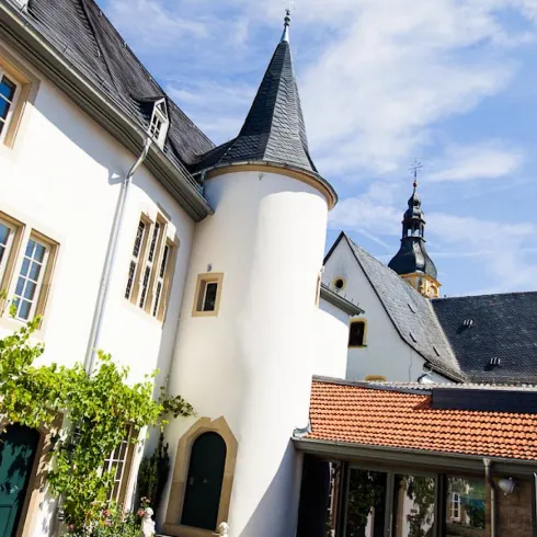 Ein historisches Gebäude mit einem spitzen Turm und bunten Fenstern. Umgeben von grünem Wein und einem klaren blauen Himmel.