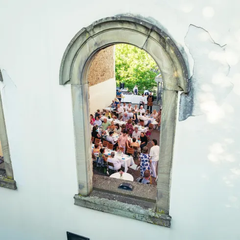 Ein festlich gedeckter Tisch mit vielen Gästen, die draußen speisen. Der Blick erfolgt durch ein großes Fenster in einer historischen Mauer.