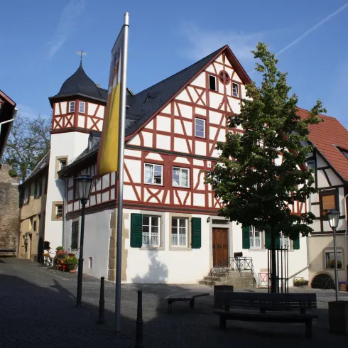 Ein historisches Fachwerkhaus mit einer weißen Fassade und roten Balken. Im Vordergrund steht ein Baum und eine ruhige Straße.