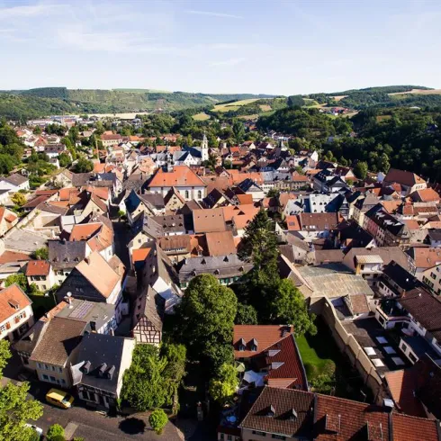 Eine malerische Stadtansicht mit vielen traditionellen Häusern und roten Dächern. Im Hintergrund sind grüne Hügel unter einem blauen Himmel zu sehen.