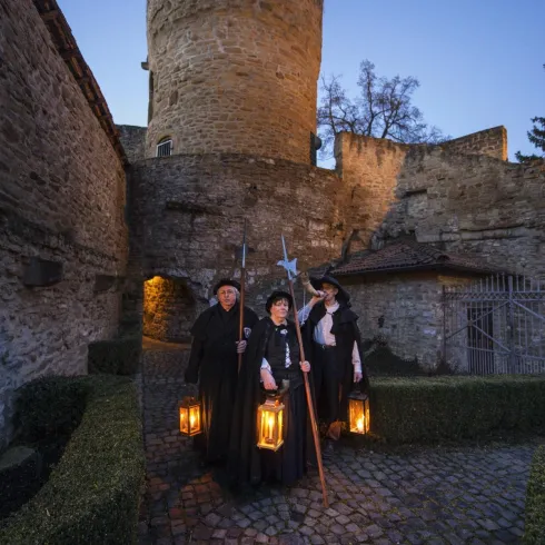 Drei Personen in historischen Kostümen stehen vor einer alten Burg. Sie halten laternen und posieren im Abendlicht.