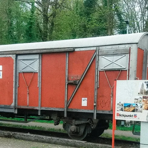 Ein roter Güterwagen steht an einem Gleis, umgeben von Bäumen. Neben dem Wagen befindet sich ein Informationsschild mit Bildern.