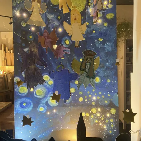 Eine farbenfrohe Wandmalerei mit Engeln und anderen Figuren vor einem nächtlichen Himmel. Im Vordergrund sind Silhouetten von Stadtgebäuden und Sternen zu sehen.