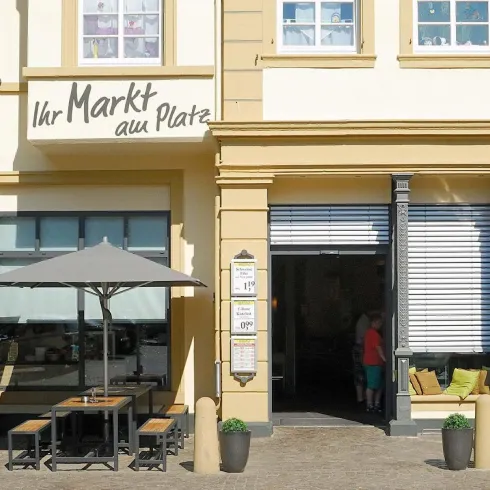 Ein einladender Markt mit Außenplätzen und Sonnenschirmen. Die Fassade ist hell und freundlich gestaltet.