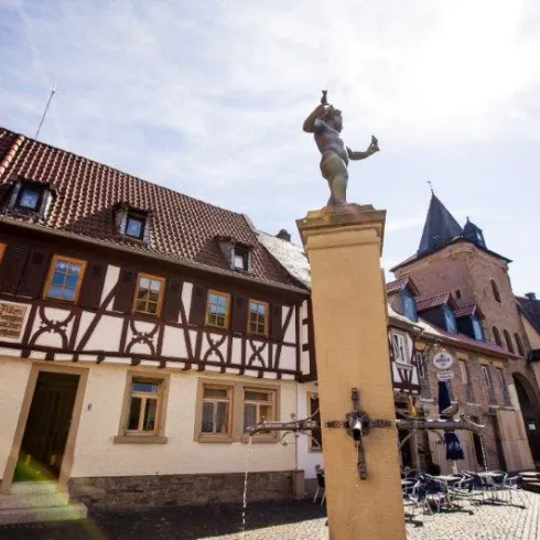 Ein malerischer Platz mit Fachwerkhäusern und einer Statue auf einem Brunnen. Im Hintergrund ist ein historisches Gebäude und ein blauer Himmel zu sehen.