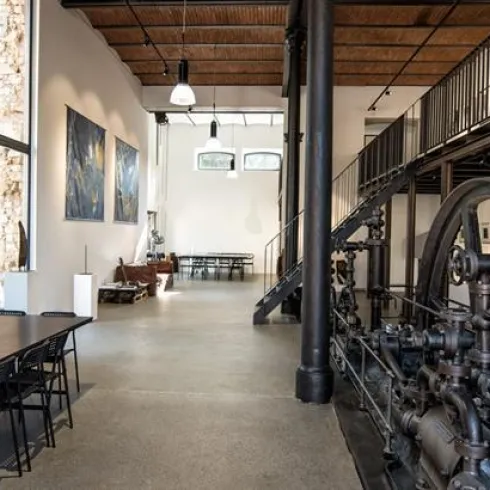 Eine moderne Galerie mit hohen Decken und großflächigen Fenstern. An den Wänden hängen zeitgenössische Kunstwerke, während im Vordergrund ein großer Tisch und ein historisches Maschinenstück zu sehen sind.