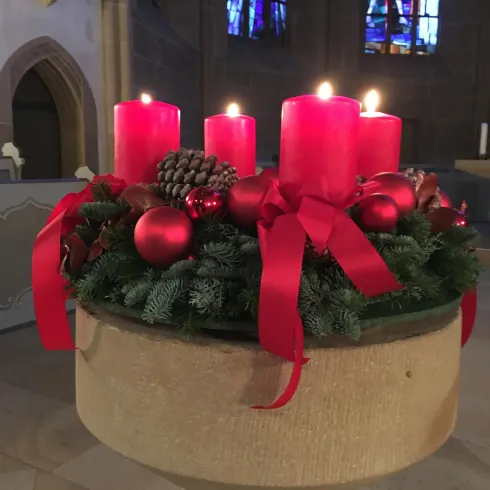 Ein Adventskranz mit vier roten Kerzen steht auf einem steinernen Podest. Er ist mit Weihnachtskugeln und roten Schleifen dekoriert.