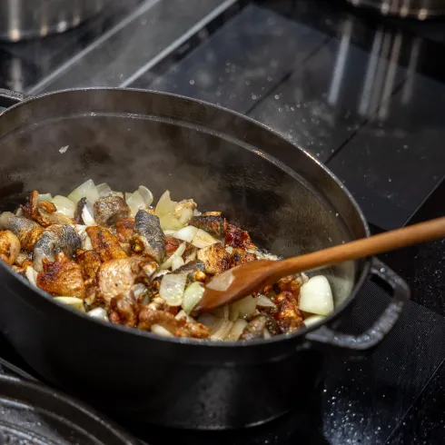 Ein großer Kochtopf mit gebratenem Gemüse und Fleisch steht auf einem modernen Kochfeld. Dampf steigt auf, während jemand mit einem Holzlöffel umrührt.