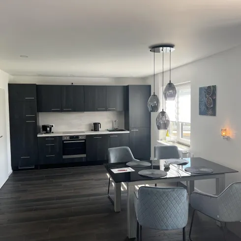 Eine moderne Küche mit dunkelgrauen Schränken und einem Essbereich. Der Raum hat große Fenster und eine elegante Beleuchtung.