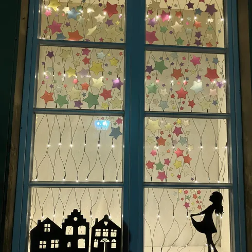 Ein Fenster mit bunten Sternen und einer Silhouette eines Mädchens. Im Vordergrund sind kleine Häuser zu sehen.