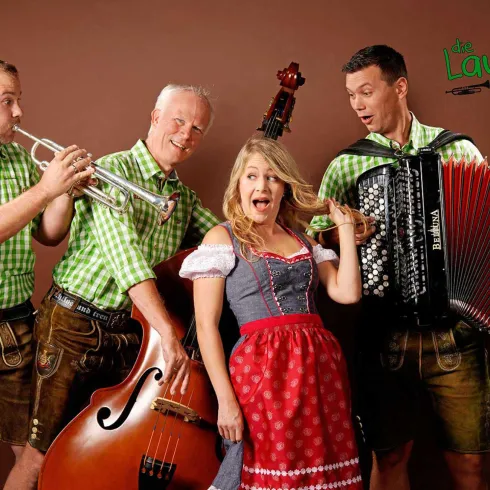 Eine Gruppe von Musikern in traditioneller Tracht spielt Instrumente. Eine Frau in einem Dirndl singt fröhlich mit drei männlichen Musikern.