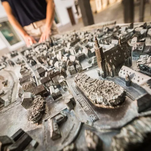 Eine detaillierte Bronzetafel zeigt ein Stadtmodell mit Gebäuden und Straßen. Im Hintergrund ist eine Person, die die Tabelle betrachtet.