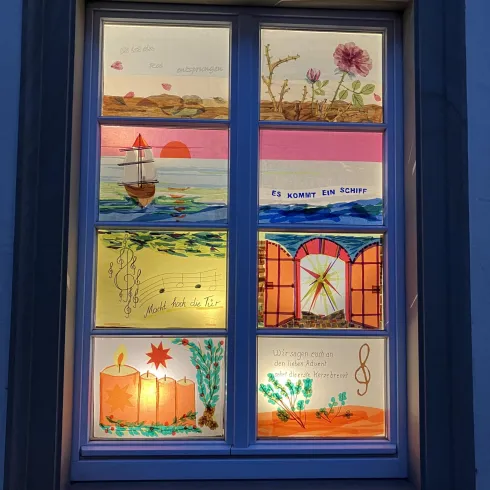 Ein Fenster mit bunten, handgemalten Bildern zeigt verschiedene Landschaften und Motive. Die Kunstwerke sind lebhaft und fröhlich, mit einem maritimen Thema und dekorativen Elementen.