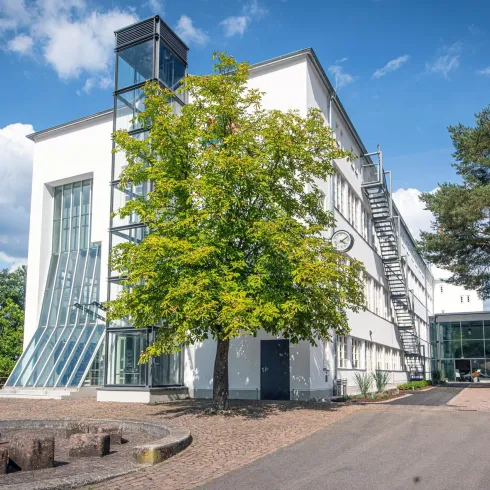 Ein modernes Gebäude mit großen Fenstern und einer Glasfassade ist umgeben von Bäumen. Der Himmel ist klar mit einigen Wolken.